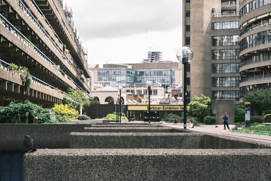 A multi-storey office building with exposed concrete balconies that have metal railings painted in dark blue, each adorned with potted plants and small shrubs. The balconies display a variety of green