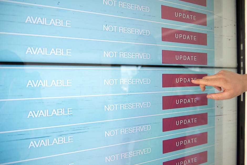 A person's hand is pointing towards a red 'Update' button on a large digital screen displaying multiple rows of schedule statuses, with several entries marked as 'Available' and 'Not Reserved.' The sc